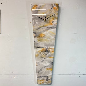 May include: A long, vertical clock with a white, gray, and gold abstract design. The clock has black hands and a gold center.