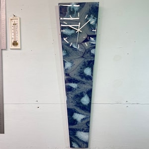 Puede incluir: Un reloj largo y estrecho, blanco y azul, con una esfera blanca y agujas negras. El reloj tiene un diseño único y abstracto con patrones en espiral.