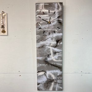May include: A long, rectangular clock with a white and gray marbled design. The clock face has black hands and black Roman numeral markings.