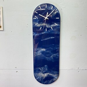 Puede incluir: Un reloj ovalado azul y blanco con una esfera blanca y agujas negras. El reloj tiene un acabado de resina y un diseño abstracto único.