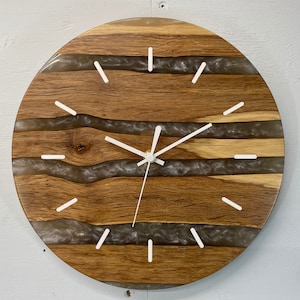 May include: A round wooden clock face with white hands and white hour markers. The wood has a natural grain and is accented with streaks of clear resin.