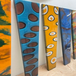 Puede incluir: Seis paneles largos, estrechos y rectangulares con diferentes diseños abstractos. Los paneles están hechos de resina y tienen un acabado brillante. Los diseños se crean con diferentes colores de resina, incluyendo azul, amarillo, naranja, verde y marrón. Los paneles están dispuestos en una fila, con el panel amarillo a la izquierda y el panel azul a la derecha.