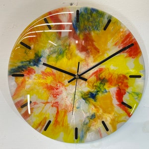 May include: A round clock with a colourful abstract design in shades of yellow, red, blue, and white. The clock face has black hour and minute hands and black markings for the hours.