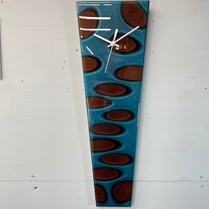 May include: A long, narrow, teal blue and brown clock with a white face and hands. The clock has a unique, abstract design with brown oval shapes on a teal blue background.