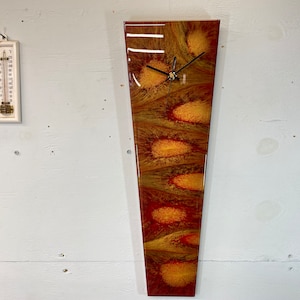 May include: A long, narrow, vertical clock with a red, orange, and gold abstract design. The clock face is white with black hands.