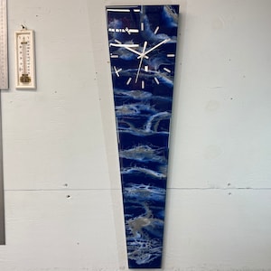 Könnte beinhalten: Eine lange, schmale, blaue und weiße Harz-Uhr mit weißem Zifferblatt und schwarzen Zeigern. Die Uhr ist an einer weißen Wand montiert.