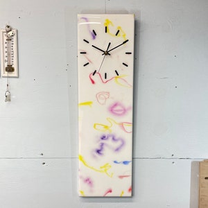 May include: A white clock with black hands and black Roman numeral markings. The clock face is embedded in a white rectangular resin panel with colorful abstract swirls in yellow, pink, and purple.