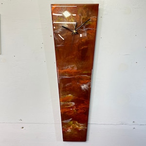 May include: A long, narrow, vertical clock with a copper-colored face and a white, abstract design. The clock has black hands and a black frame.