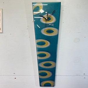 Puede incluir: Un reloj abstracto de color verde azulado y amarillo con fondo blanco. La esfera del reloj es un rectángulo vertical con una serie de círculos amarillos sobre un fondo verde azulado.