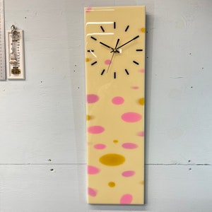 May include: A long, rectangular clock with a white background and pink and yellow abstract shapes. The clock face is black with white hands and black numbers.