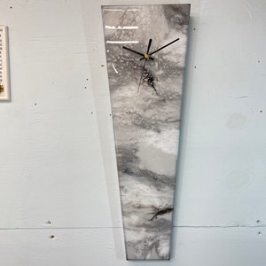 Peut inclure: Une horloge en résine abstraite blanche et grise avec des aiguilles noires. L'horloge a la forme d'un long rectangle fin.