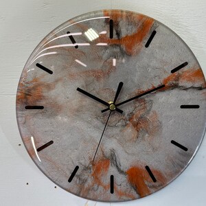 May include: A round, grey and orange resin clock with black hands and black Roman numeral markings.