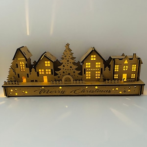 Wooden Christmas light up decoration featuring a village scene with tree and houses. Laser cut wood is hand assembled with LED lights.