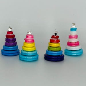 May include: Four colourful button Christmas tree decorations. Each tree is made of stacked buttons in a rainbow pattern, with a silver star on top. The trees are arranged in a row on a white surface.
