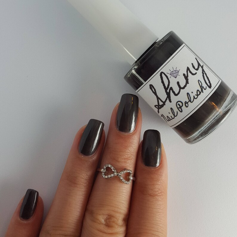 May include: A hand wearing a dark gray nail polish with an infinity ring. The hand is holding a bottle of nail polish labeled "Shiny Nail Polish".