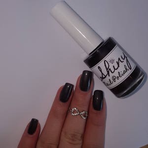 May include: A bottle of dark gray nail polish with a white label that says "Shiny Nail Polish". A hand with dark gray nail polish is shown wearing a silver infinity ring.