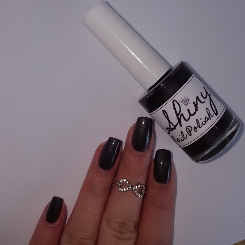 May include: A bottle of dark gray nail polish with a white label that says "Shiny Nail Polish". A hand with dark gray nail polish is shown wearing a silver infinity ring.