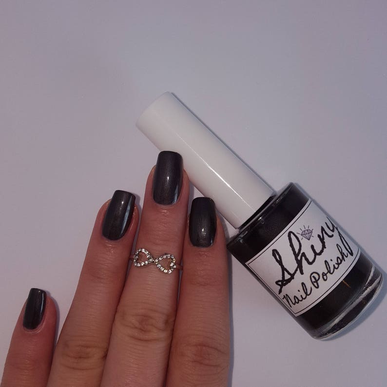 May include: A bottle of dark gray nail polish with a white cap and the text "Shiny Nail Polish" on the label. The bottle is sitting on a white surface. A hand with dark gray nail polish is holding an infinity ring.