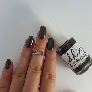 May include: A dark gray nail polish bottle with a white label that says "Shiny Nail Polish". A hand is wearing dark gray nail polish and an infinity ring.
