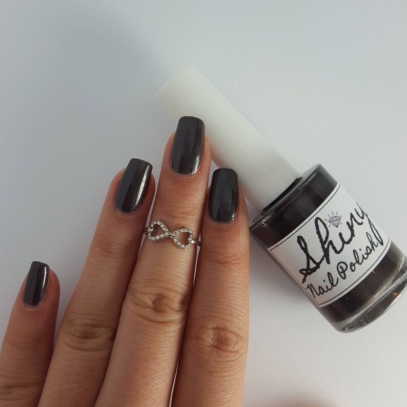 May include: A dark gray nail polish bottle with a white label that says "Shiny Nail Polish". A hand is wearing dark gray nail polish and an infinity ring.