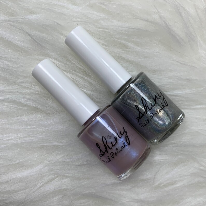 May include: Two bottles of nail polish, one is a light purple with a shimmer and the other is a silver with a shimmer. Both bottles have the text "Shiny Nail Polish" on the label.