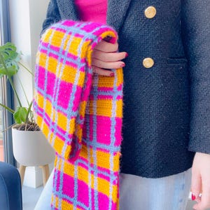 Op de afbeelding: Een kleurrijke, met de hand gehaakte sjaal met een levendig ruitpatroon in roze, geel en lichtblauw. De sjaal wordt getoond tegen een donkere blazer met gouden knopen en een lichtblauwe jeans. Het patroon van de sjaal is opvallend.