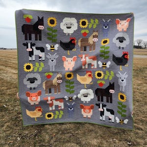May include: A gray quilt featuring various farm animals, including cows, pigs, sheep, and chickens, along with sunflowers and bees. The quilt has a green border and is held up against a cloudy sky.