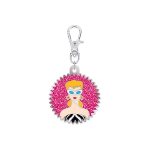 May include: A silver-toned keychain with a round, pink glittery pendant featuring a cartoon woman with blonde hair, blue sunglasses, and a black and white top. The pendant has a silver border and a lobster clasp.