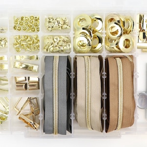 May include: A clear plastic storage box filled with gold-colored metal zipper pulls, snaps, eyelets, and other hardware. The box also includes three rolls of zipper tape in gray, beige, and brown.