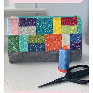 May include: A colorful patchwork fabric pouch with a zipper closure. The pouch is 7 inches by 11 inches by 2 inches. The pouch is made with a variety of fabrics in shades of pink, red, yellow, green, blue, and purple. The pouch is sitting on a wooden surface with a spool of blue thread and a pair of scissors.