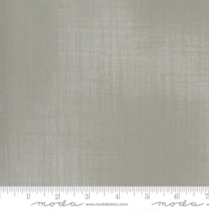 May include: A close-up of a light gray fabric with a subtle textured pattern. The fabric is measured in inches with the text "moda" printed on the ruler.