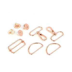 May include: A set of rose gold metal hardware for bags and purses. The set includes two D-rings, two swivel clasps, a buckle, two snap fasteners, and a lobster clasp.