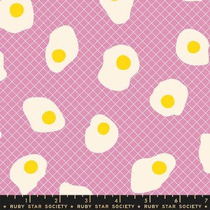 May include: Fabric with a repeating pattern of fried eggs on a pink grid background. The eggs have yellow yolks and off-white whites. The fabric is from Ruby Star Society.