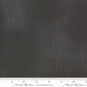 May include: A close-up of a dark gray fabric with a subtle texture. The fabric is measured with a ruler in inches, with the word "moda" printed on the ruler.