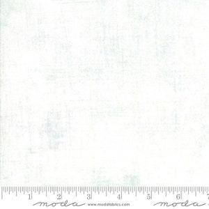 May include: A white fabric with a subtle, light blue, textured pattern. The fabric is marked with a ruler in inches and the word "moda" appears twice on the ruler.