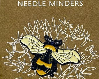 May include: A bee-shaped needle minder with yellow and black enamel, set against a gold background with white floral accents. Text reads "MIND the pollinators" and "NEEDLE MINDERS". Signed "Kate Wyatt FINE ART".