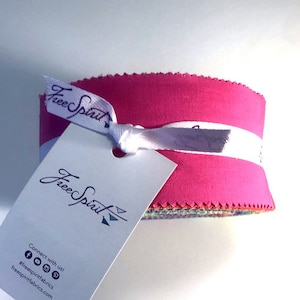 May include: A pink fabric bundle with a white label that says "Free Spirit" and a smaller white label with the same text and a triangle design. The bundle is tied with a white ribbon.
