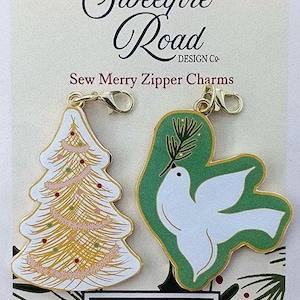May include: Two Christmas-themed zipper charms, one featuring a white Christmas tree with gold and pink decorations and the other a white dove with a green olive branch, both with gold accents. The charms are attached to gold zipper pulls. The packaging features the text "Sweetfire Road Design Co. Sew Merry Zipper Charms" and the Moda Fabrics + Supplies logo.