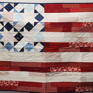 May include: A handmade quilt featuring the design of the American flag. The quilt showcases a patchwork of red and white stripes, with a blue square in the upper left corner filled with white and blue diamond and star patterns. The quilt is bordered with a dark blue fabric.
