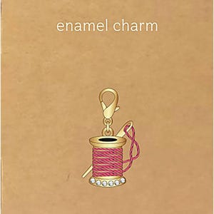 May include: A gold-toned enamel charm shaped like a spool of pink thread with a needle, featuring clear rhinestones at the base and a lobster clasp. The charm is displayed on a tan card with the text enamel charm and JEWELRY MADE BY ME.