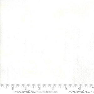 May include: White fabric with a subtle texture. The fabric is marked with a ruler and the word "moda" appears twice on the fabric.