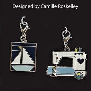 May include: Two metal charms with silver clasps. One charm is a sailboat design in navy, white, and light blue. The other is a sewing machine in white and light blue. The text "Designed by Camille Roskelley" is at the top.
