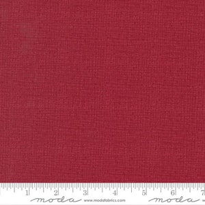 May include: A solid red fabric with a subtle textured pattern. The fabric is marked with a ruler in inches and the word "moda" appears twice on the ruler.