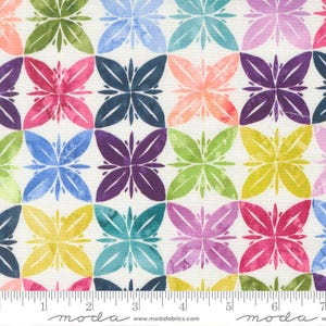 May include: A fabric with a repeating pattern of colorful, stylized floral shapes in shades of pink, purple, blue, green, and yellow. The flowers are arranged in a grid on a cream-colored background. The fabric has a ruler along the bottom with the text "moda" and "www.modafabrics.com".