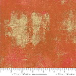 May include: A close-up of a textured fabric with a gold metallic sheen. The fabric has a mottled orange and red color with a subtle pink hue. The fabric is measured with a ruler that reads "moda" and includes the website address "www.modafabrics.com".
