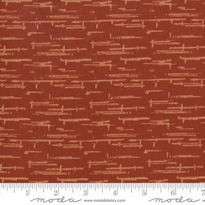 May include: A rust-colored fabric featuring a repeating pattern of horizontal, dashed, and textured light beige lines. A white ruler is visible at the bottom edge, showing measurements in inches, with the text moda and www.modafabrics.com printed.