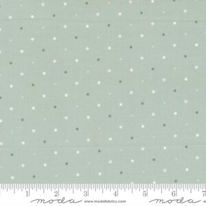 May include: A light sage green fabric featuring a repeating pattern of small white polka dots. A white ruler is visible at the bottom edge, showing a scale of 7 inches, with the text "moda" and "www.modafabrics.com" printed along the bottom.