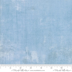 May include: A light blue fabric with a subtle textured pattern. The fabric has a slightly distressed look, with faded areas and a hint of white throughout. The fabric is marked with a ruler and the word "moda" in a handwritten font.