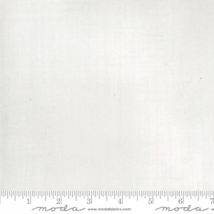 May include: White fabric with a subtle texture. The fabric is measured with a ruler that has the word "moda" printed on it.