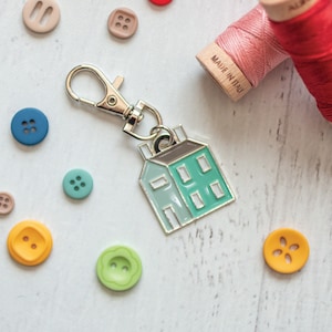 May include: A silver keychain with a teal and gray house charm. The charm has a small silver loop at the top and is attached to a silver split ring. The keychain is surrounded by colorful buttons.